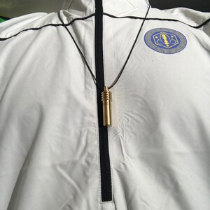 May include: A gold-colored cylindrical pendant necklace with a black cord. The pendant has a ribbed top and a smooth, elongated body. The necklace hangs against a white jacket with a black zipper and trim. A blue and gold emblem is visible on the jacket.