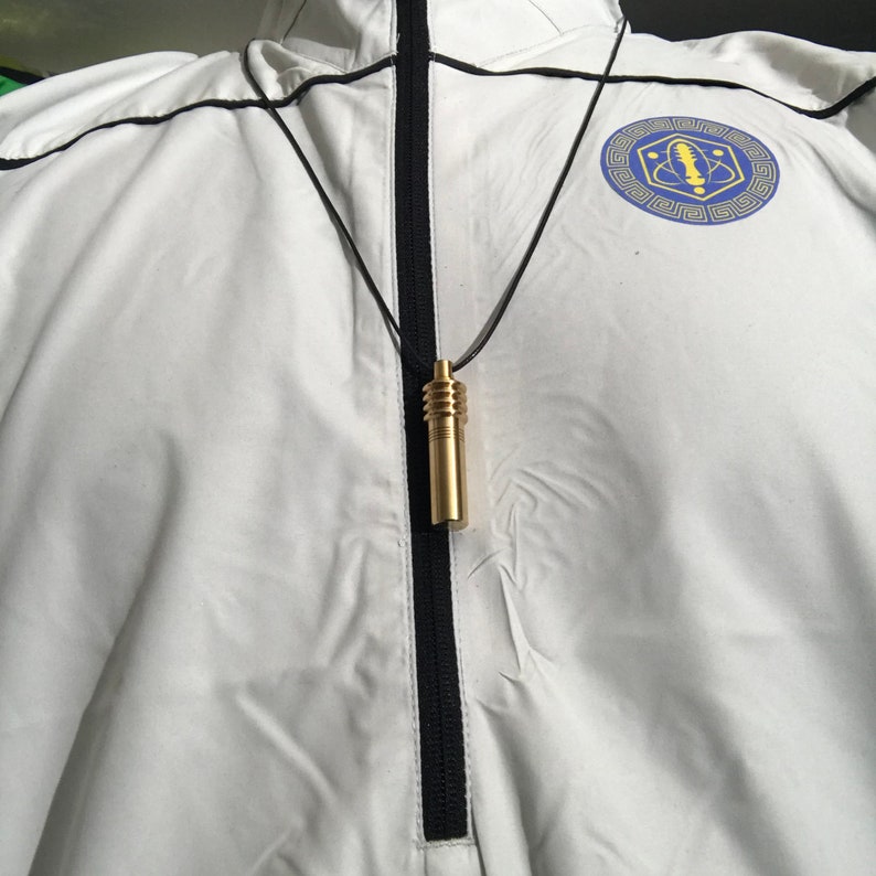 May include: A gold-colored cylindrical pendant necklace with a black cord. The pendant has a ribbed top and a smooth, elongated body. The necklace hangs against a white jacket with a black zipper and trim. A blue and gold emblem is visible on the jacket.