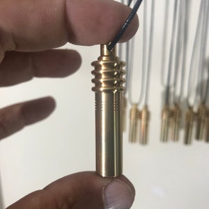 May include: A gold-colored metal pendant necklace with a cylindrical design. The pendant is held by a black cord. The pendant has a ridged top and a series of horizontal lines near the bottom. Several similar necklaces are visible in the background.
