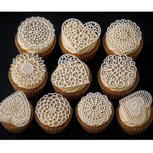 Puede incluir: Vista cenital de once cupcakes con glaseado blanco y adornos de encaje blanco intrincados en varias formas, incluyendo corazones, círculos y un girasol. Los cupcakes están sobre una superficie negra.