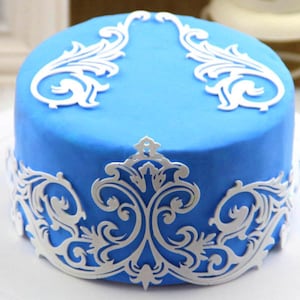 Puede incluir: Un pastel azul decorado con encaje de fondant blanco. El encaje tiene un delicado patrón floral y se coloca alrededor de la parte inferior y superior del pastel.