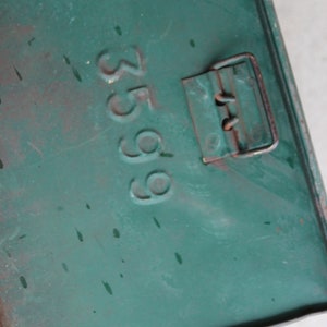 May include: Close-up of a weathered, teal-colored metal box. The number "3599" is stamped on the surface. A rusty metal clasp is visible on the right side. The box has paint splatters and signs of wear.