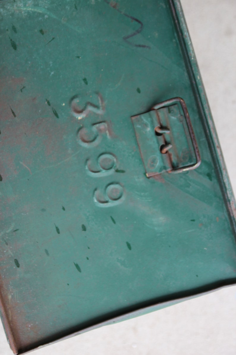 May include: Close-up of a weathered, teal-colored metal box. The number "3599" is stamped on the surface. A rusty metal clasp is visible on the right side. The box has paint splatters and signs of wear.