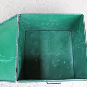 May include: An open, rectangular green metal storage box with a hinged lid. The box has a handle on the front and is set against a light-colored surface. The interior of the box is also green.