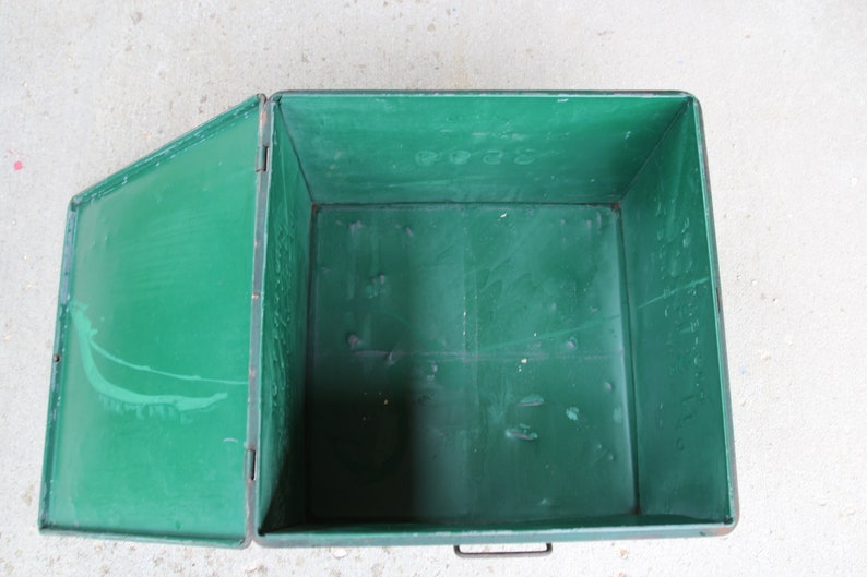 May include: An open, rectangular green metal storage box with a hinged lid. The box has a handle on the front and is set against a light-colored surface. The interior of the box is also green.
