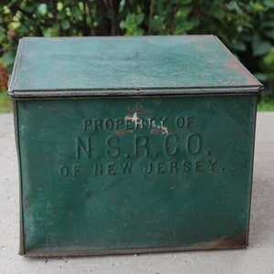 May include: A vintage, square, green metal box with a hinged lid. The box has the text "PROPERTY OF N.S.R.CO. OF NEW JERSEY" embossed on the front. The paint is distressed, showing signs of age and wear.