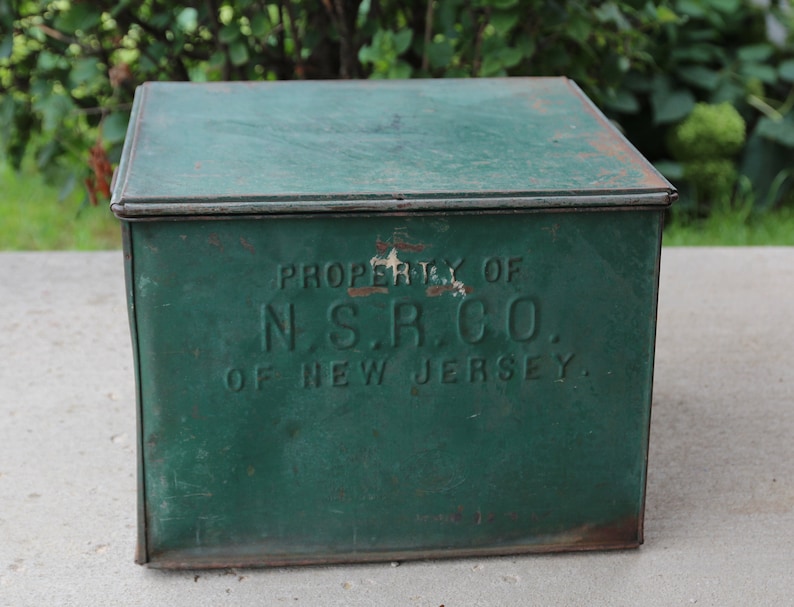May include: A vintage, square, green metal box with a hinged lid. The box has the text "PROPERTY OF N.S.R.CO. OF NEW JERSEY" embossed on the front. The paint is distressed, showing signs of age and wear.