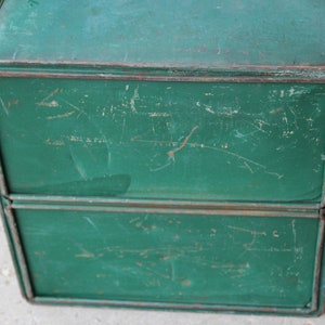 May include: A vintage, two-tiered metal box with a distressed, green painted finish. The box has a rectangular shape with visible wear and tear, including scratches and rust, giving it a rustic appearance. The edges are reinforced with metal trim.
