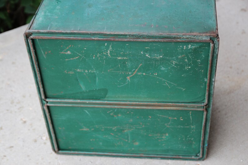 May include: A vintage, two-tiered metal box with a distressed, green painted finish. The box has a rectangular shape with visible wear and tear, including scratches and rust, giving it a rustic appearance. The edges are reinforced with metal trim.