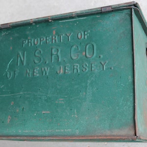 May include: A vintage green metal box with a hinged lid and a latch. The box has the text "PROPERTY OF N.S.R. CO. OF NEW JERSEY" embossed on the front. The box shows signs of wear and tear, with some rust and scratches.
