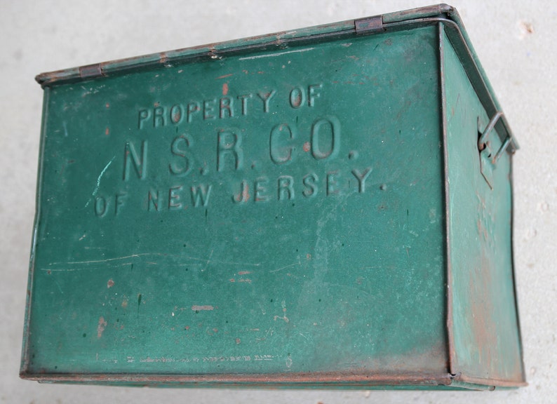 May include: A vintage green metal box with a hinged lid and a latch. The box has the text "PROPERTY OF N.S.R. CO. OF NEW JERSEY" embossed on the front. The box shows signs of wear and tear, with some rust and scratches.