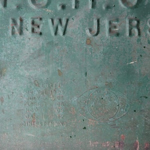 May include: Close-up of a weathered, green metal surface with embossed text. The words "OF NEW JERSEY" are visible. Additional text includes dates and the word "PATENTED". A circular emblem is also present.