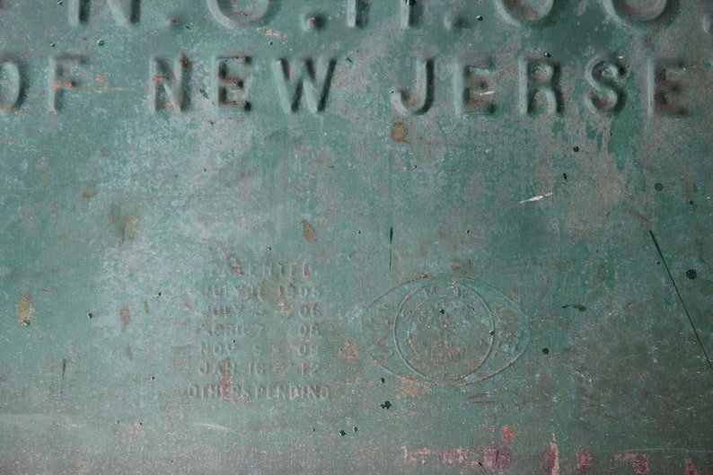May include: Close-up of a weathered, green metal surface with embossed text. The words "OF NEW JERSEY" are visible. Additional text includes dates and the word "PATENTED". A circular emblem is also present.