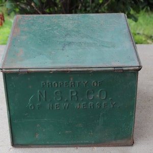May include: A vintage green metal box with a hinged lid. The box has visible rust and the text "PROPERTY OF N.S.R. CO. OF NEW JERSEY" is embossed on the front. The box is likely used for storage.