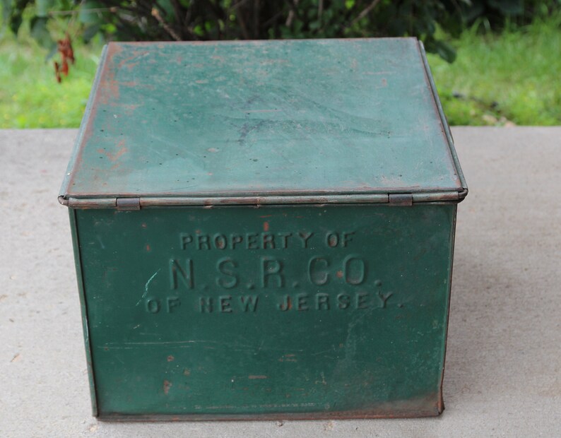May include: A vintage green metal box with a hinged lid. The box has visible rust and the text "PROPERTY OF N.S.R. CO. OF NEW JERSEY" is embossed on the front. The box is likely used for storage.