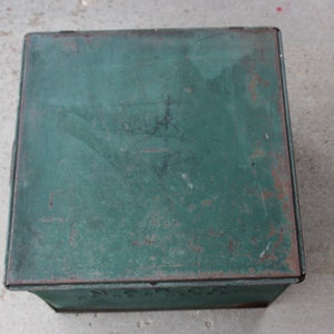 May include: A vintage, square metal box with a hinged lid. The box is painted a distressed green color with visible rust spots, giving it a rustic appearance. The box is likely used for storage.
