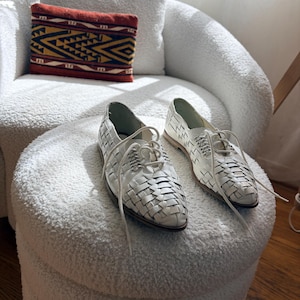 May include: A pair of white leather woven lace-up shoes with a pointed toe. The shoes are displayed on a white, textured ottoman. A decorative pillow with a geometric pattern in red, yellow, and black rests on a white armchair in the background.