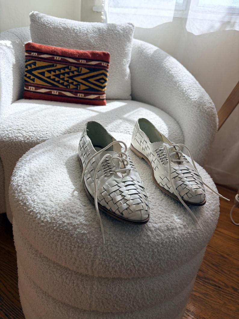 May include: A pair of white leather woven lace-up shoes with a pointed toe. The shoes are displayed on a white, textured ottoman. A decorative pillow with a geometric pattern in red, yellow, and black rests on a white armchair in the background.