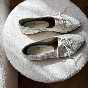 May include: A pair of white woven leather shoes with laces, displayed on a white, fluffy, round ottoman. The shoes have a low heel and a pointed toe. The insoles are visible, with text that reads "NATURALES".