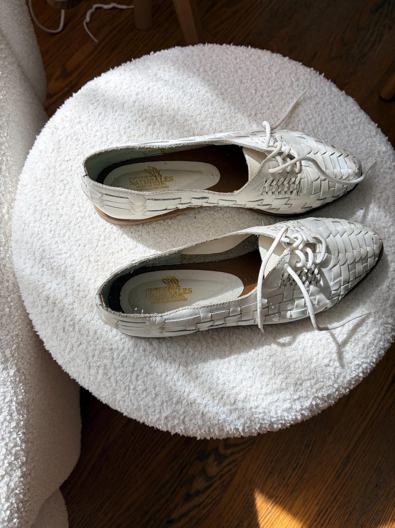 May include: A pair of white woven leather shoes with laces, displayed on a white, fluffy, round ottoman. The shoes have a low heel and a pointed toe. The insoles are visible, with text that reads "NATURALES".