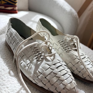 May include: A pair of white leather woven shoes with laces. The shoes have a low profile and a brown sole. The woven design creates a textured appearance. The shoes are displayed on a white surface.