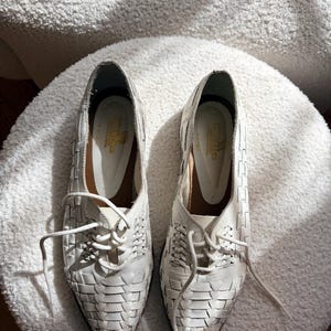 May include: A pair of white leather woven lace-up shoes. The shoes have a pointed toe and a low heel. The woven design covers the entire shoe, with laces that extend from the top of the shoe. The shoes are placed on a white, textured surface.
