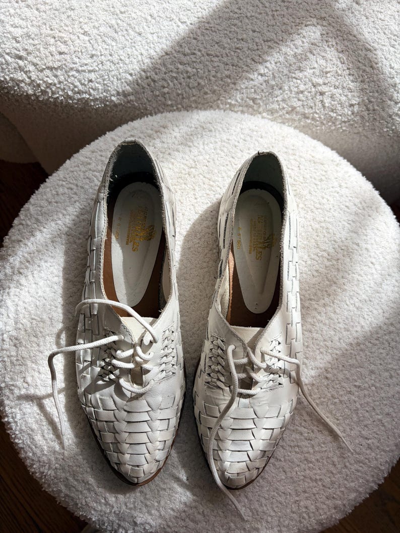 May include: A pair of white leather woven lace-up shoes. The shoes have a pointed toe and a low heel. The woven design covers the entire shoe, with laces that extend from the top of the shoe. The shoes are placed on a white, textured surface.