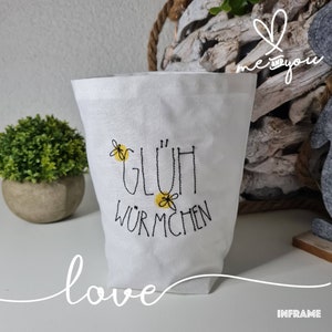 Può includere: Borsa di tela bianca con ricamo nero che dice "Glühwürmchen" con fiori ricamati gialli. La borsa è su una superficie bianca con la parola "love" scritta in corsivo nero.