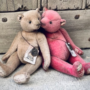 Könnte beinhalten: Zwei Vintage-Teddybären, einer beige und einer rosa, sitzen nebeneinander. Der beige Bär hat ein Etikett mit der Nummer "077" darauf. Der rosa Bär hat ein Etikett mit einem kleinen Design.
