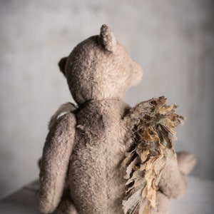 Peut inclure: Un ours en peluche de style vintage, vu de dos, avec des ailes déchirées. L'ours est de couleur marron clair et est assis sur une surface claire. Les ailes ont un aspect texturé et vieilli.