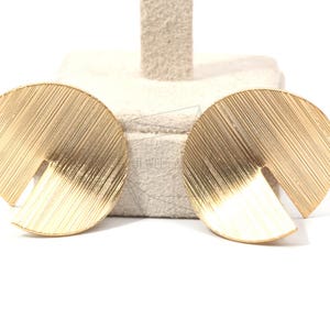 Può includere: Un paio di orecchini dorati con un design geometrico semicircolare. Gli orecchini hanno una superficie testurizzata e striata.