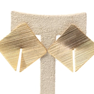 Può includere: Un paio di orecchini quadrati color oro con un design lineare strutturato. Ogni orecchino presenta un taglio diagonale su un lato, creando una forma geometrica unica. Gli orecchini sono esposti su un espositore per orecchini dai toni neutri.