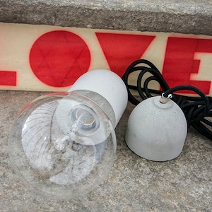 Può includere: Una lampada a sospensione in cemento grigio con una lampadina in vetro trasparente e un cavo nero. La luce è appesa a un apparecchio a soffitto in cemento grigio.