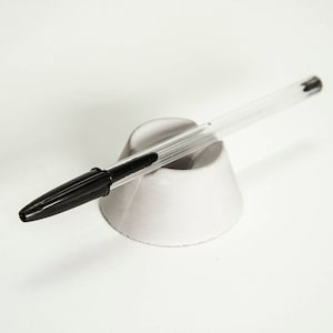 May include: A clear pen with a black ink tip is resting in a white pen holder.