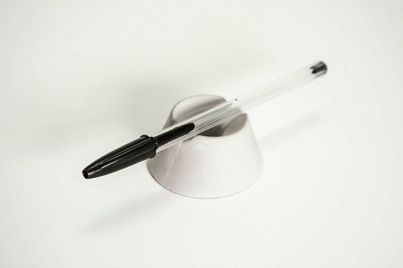 May include: A clear pen with a black ink tip is resting in a white pen holder.