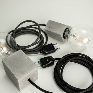 Puede incluir: Dos lámparas colgantes de hormigón gris con cables eléctricos negros y bombillas de vidrio transparente. Cada lámpara tiene un enchufe negro de dos clavijas.
