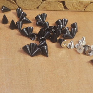 Puede incluir: Una colección de tachuelas piramidales de metal negro y tornillos plateados. Las tachuelas tienen una punta puntiaguda y una base plana. Los tornillos tienen cabeza de estrella y una arandela plana.