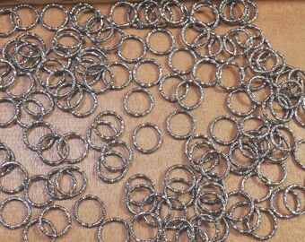 May include: A large quantity of silver jump rings, a type of jewellery finding, are scattered on a brown surface. The rings are all the same size and are made of a shiny metal.
