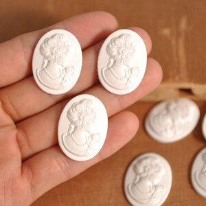 May include: White oval cameo pendants featuring a classical woman's profile. The pendants have a raised relief design with intricate details of the woman's hair, necklace, and clothing. Several cameos are visible.
