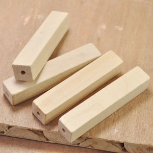 Puede incluir: Varios bloques de madera rectangulares sin terminar, de color madera claro y natural. Cada bloque tiene un extremo cuadrado con un agujero perforado. Los bloques están dispuestos sobre una superficie de madera marrón claro.