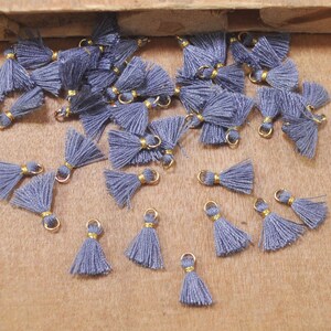 Puede incluir: Una colección de abalorios de borla gris con anillos dorados. Las borlas están hechas de hilo suave y esponjoso y son perfectas para agregar un toque de fantasía a joyas, bolsos u otras manualidades.