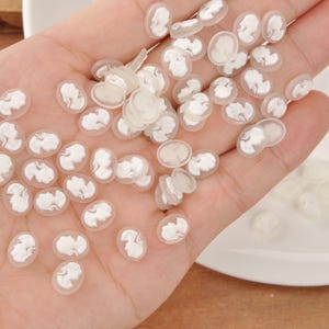 May include: A collection of small, oval-shaped cameo beads. Each bead features a white profile design set against a translucent, off-white background. The beads are scattered across a surface, showcasing their delicate details.