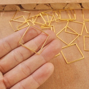 Puede incluir: Una mano sosteniendo un anillo de salto cuadrado dorado. Hay muchos más anillos de salto dorados esparcidos sobre una superficie de madera.