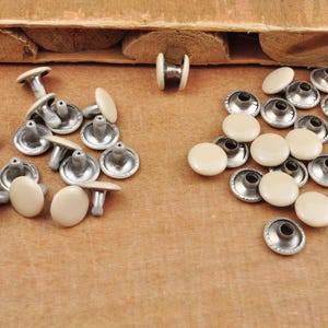 May include: A collection of off-white metal rivets with a silver finish. The rivets have a flat, round head and a post with a small hole. The rivets are arranged in two groups on a wooden surface.
