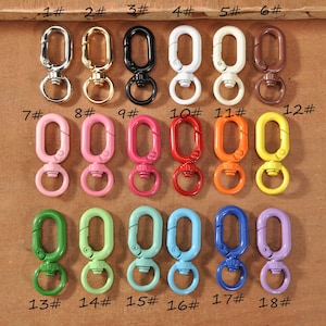 Puede incluir: Una colección de dieciocho cierres giratorios de metal en varios colores, incluyendo plata, oro, negro, blanco, marrón, rosa, rojo, naranja, amarillo, verde, verde claro, turquesa, azul y morado. Cada cierre tiene un anillo y un cierre de mosquetón.