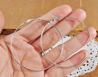 May include: A pair of silver hoop earrings. The earrings are large and circular, and they are shown on a hand.