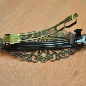 May include: A gold-toned hair clip with a decorative, intricate, floral design. The clip has a spring-loaded mechanism for holding hair.