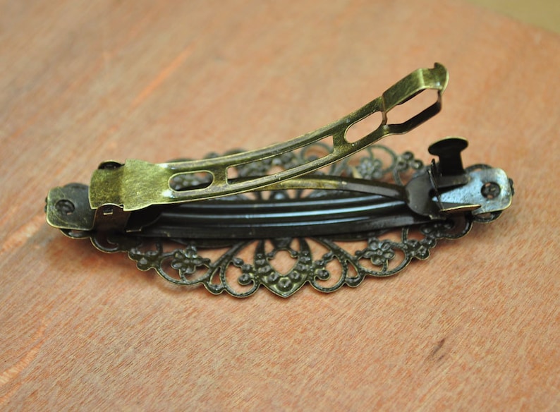 May include: A gold-toned hair clip with a decorative, intricate, floral design. The clip has a spring-loaded mechanism for holding hair.