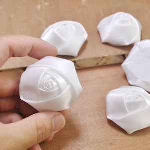 May include: Five white satin fabric rose flower embellishments. The roses are approximately 1.5 inches in diameter.