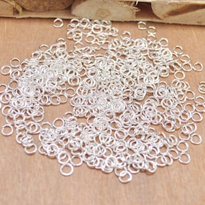 May include: A large pile of silver jump rings, a type of jewellery finding, scattered on a wooden surface.